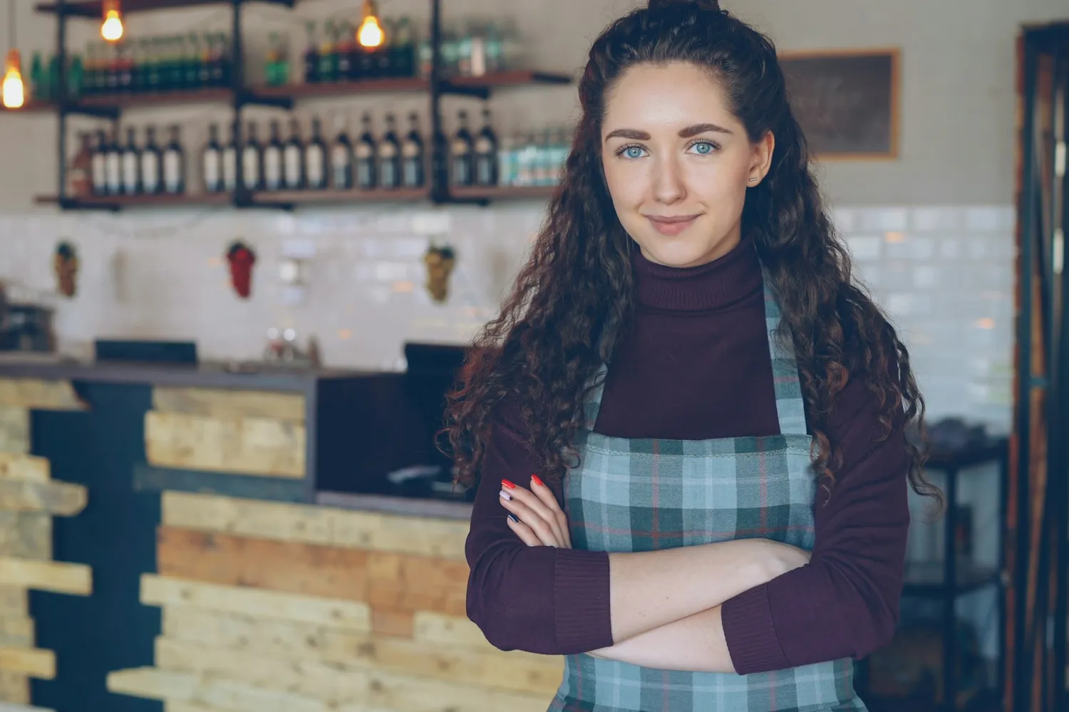 A smiling coffee shop owner in an apron, arms folded, in front of her counter