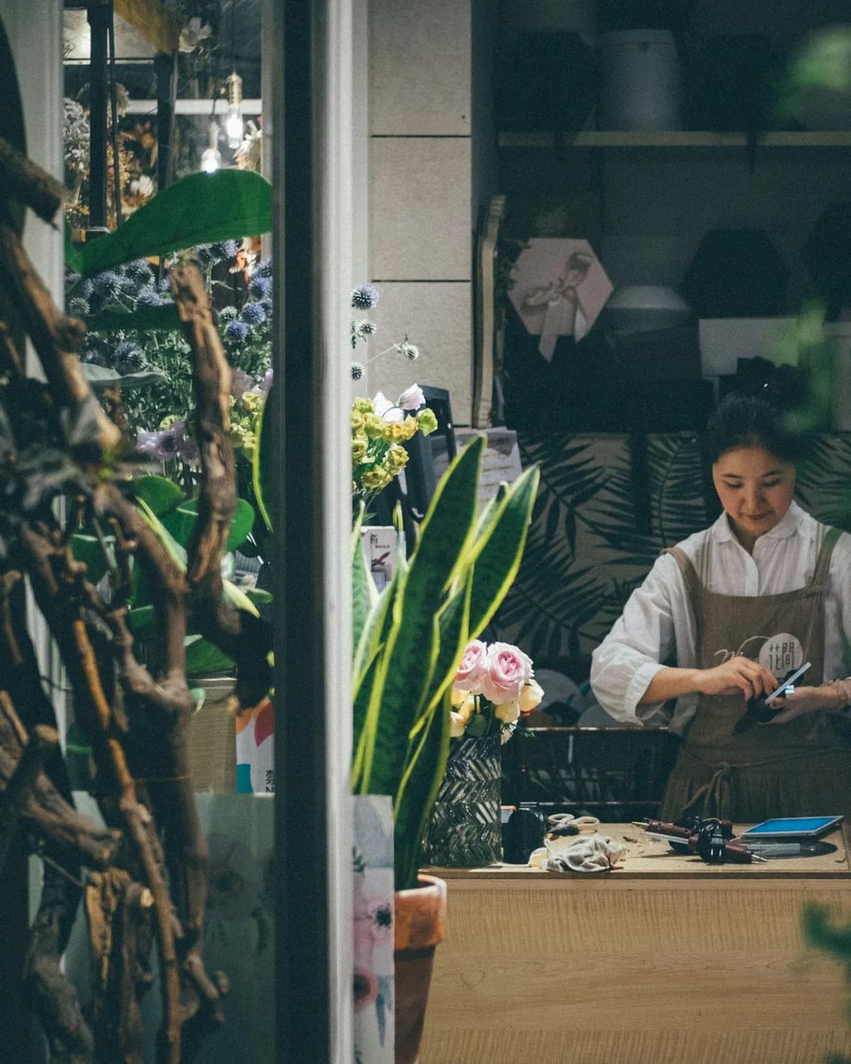 A florist preparing an arrangement at her workbench, seen through plants in the shop doorway