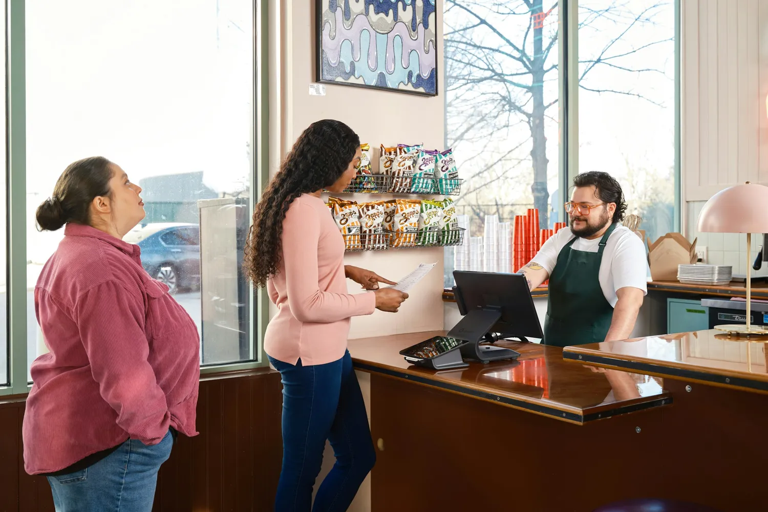 Customers ordering at a sunny café counter from a cashier beside a point-of-sale screen